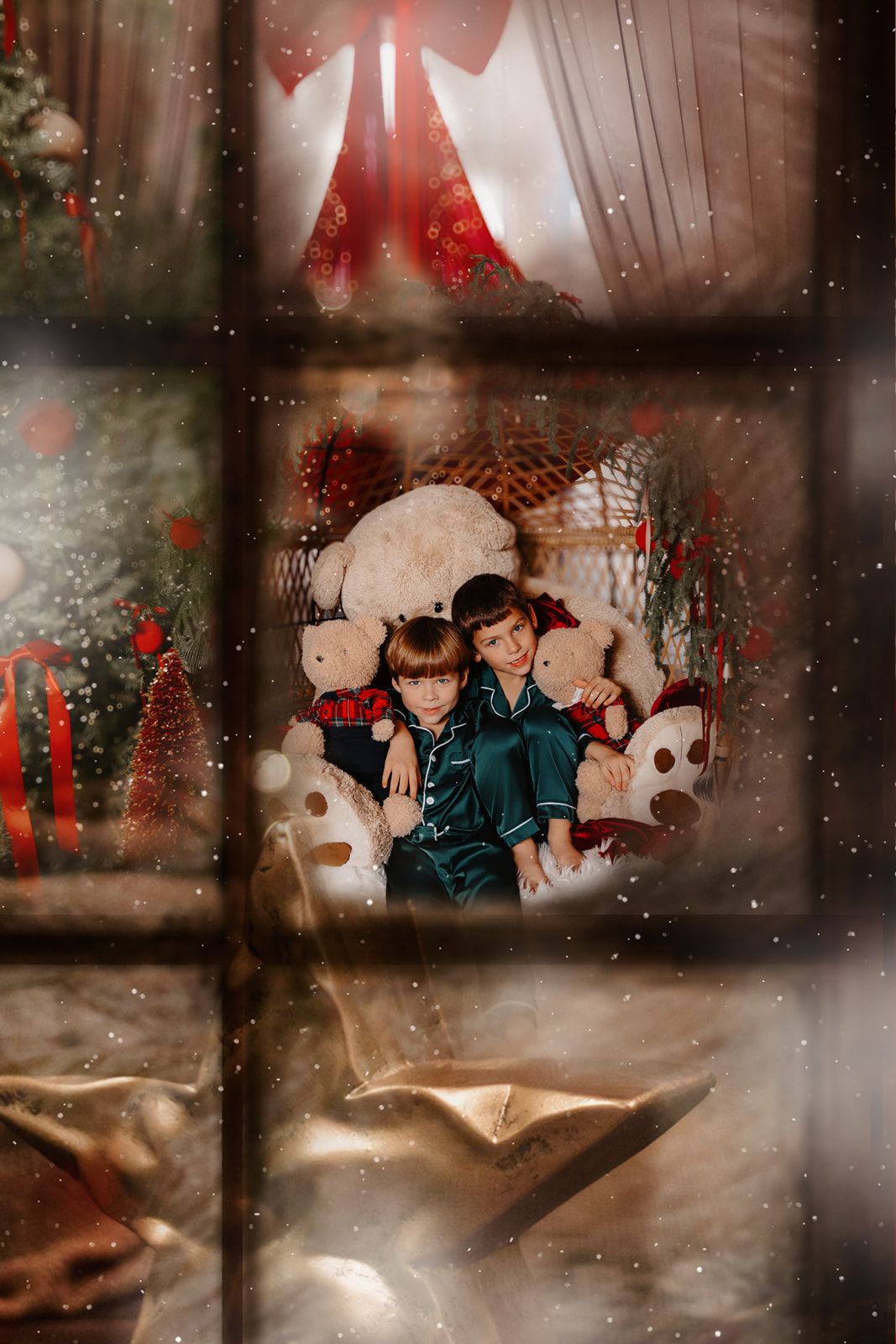 Two children in green pajamas cuddle among teddy bears in a festive Christmas decorated room, viewed through a snowy window.