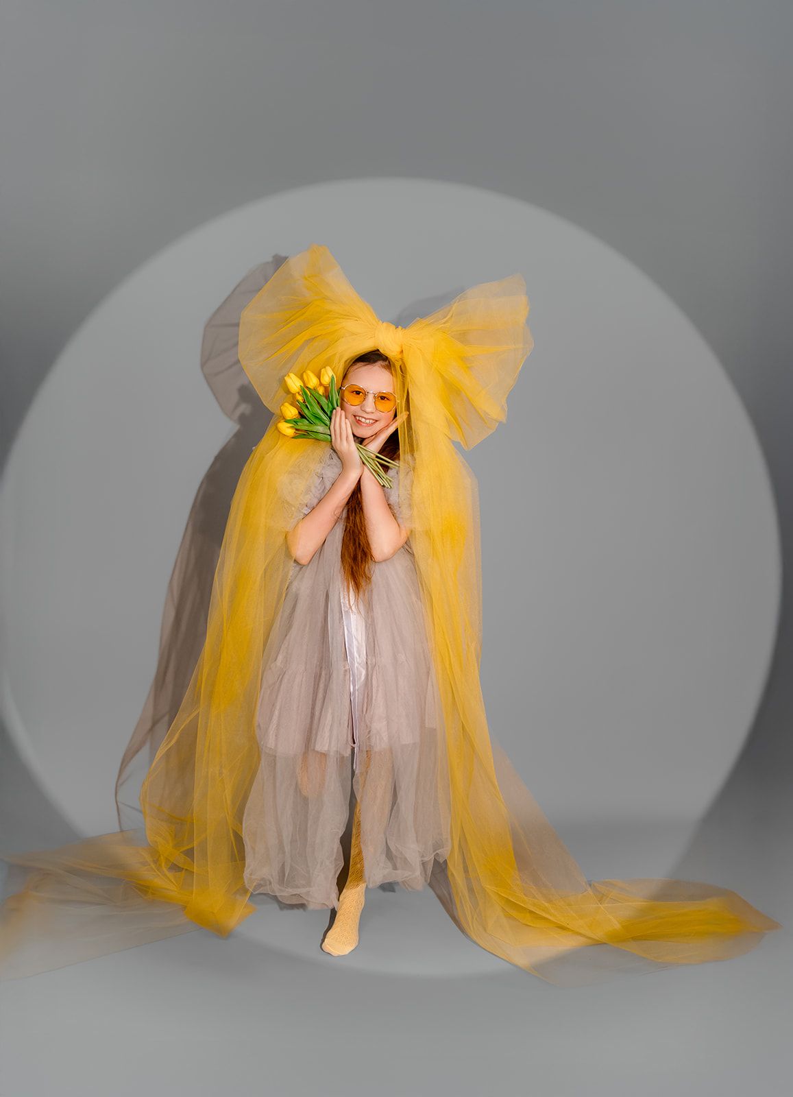 Young woman in yellow tulle gown holding tulips, wearing yellow sunglasses, posing in studio spotlight.