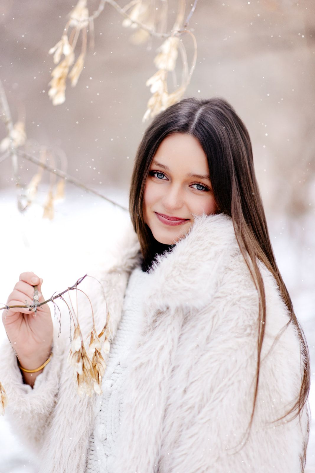Pretty brunette woman in white fluffy coat smiling while holding snowy tree branch during light snowfall outdoors.