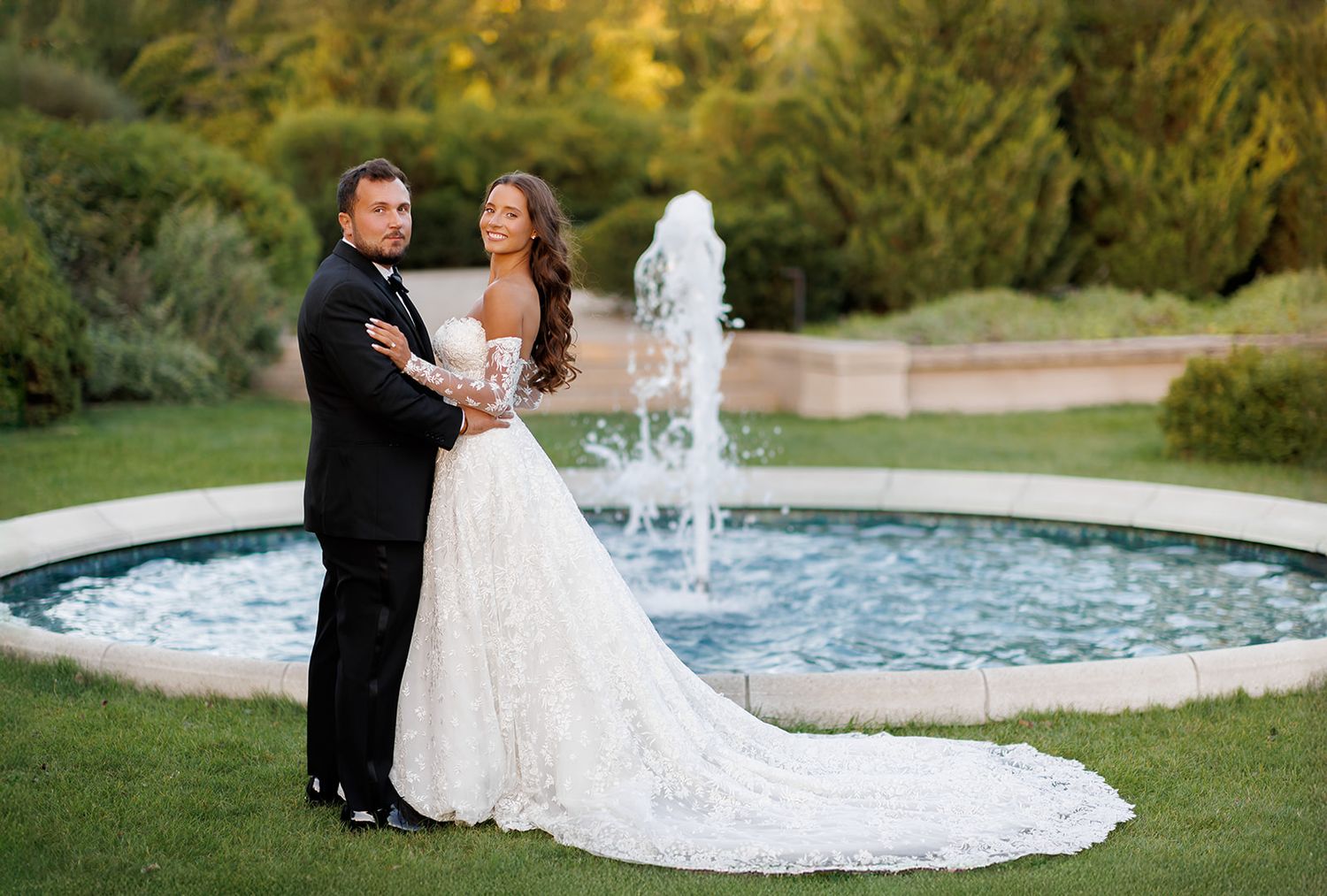 Bride and groom embrace by circular fountain in lush garden at golden hour wedding portrait.