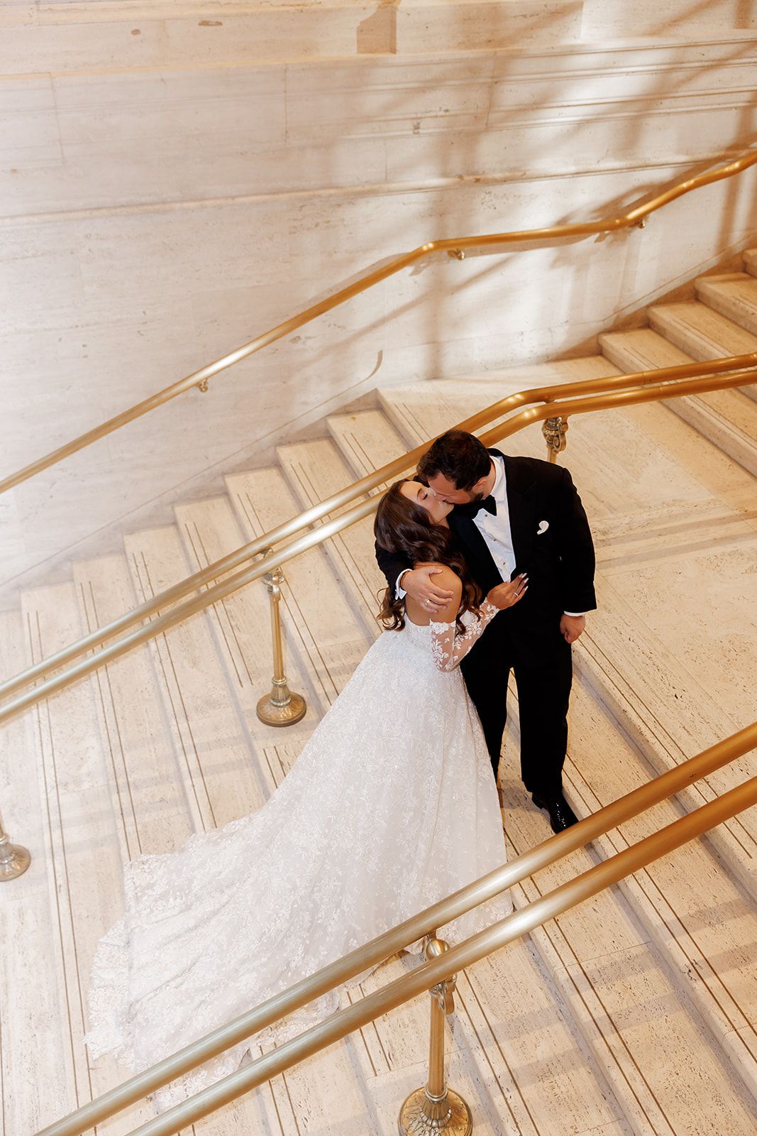 Newlyweds share a romantic kiss on elegant marble staircase with gold railings during wedding photos.