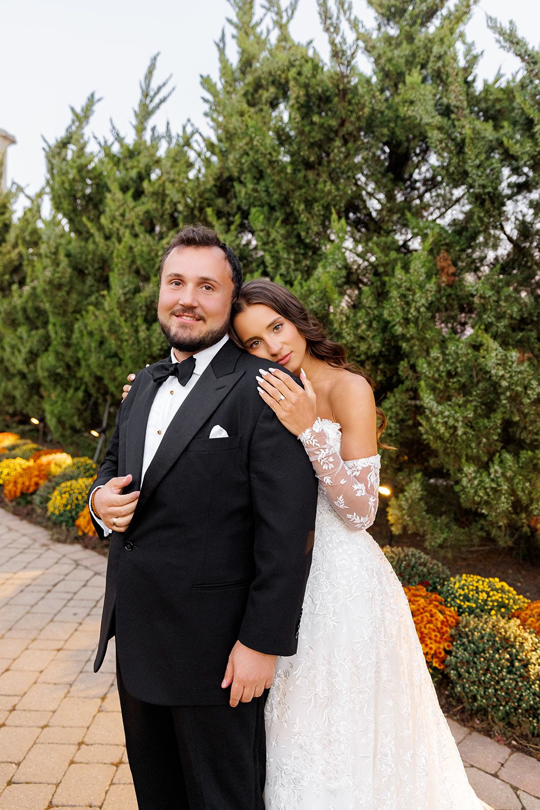 A bride in lace gown leans on groom in black tuxedo, posing before evergreen trees and autumn flowers.