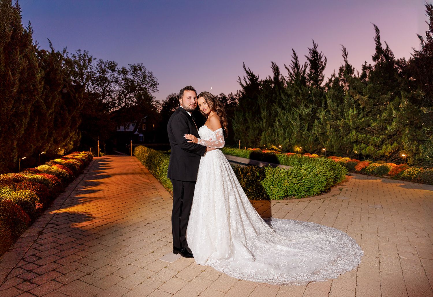 Bride and groom embrace on illuminated brick pathway at twilight, surrounded by manicured hedges and warm garden lights.