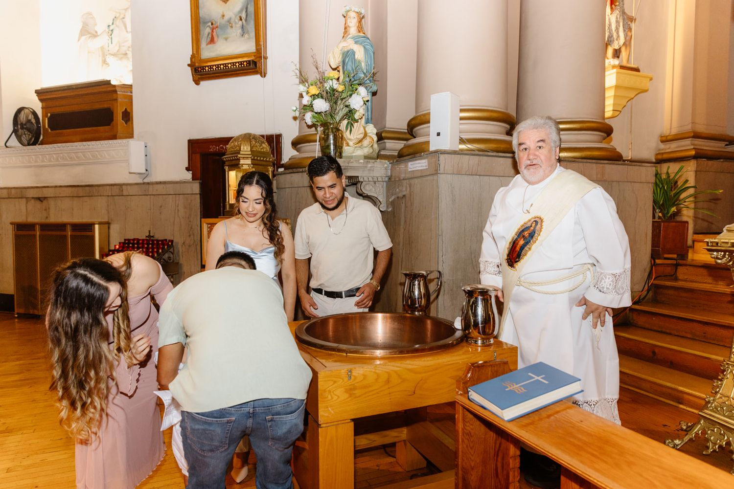 Priest and family members gathered around baptismal font during Catholic christening ceremony.