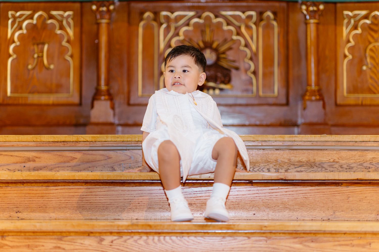 A toddler in white outfit and socks sits on wooden church steps, looking thoughtfully to the side.