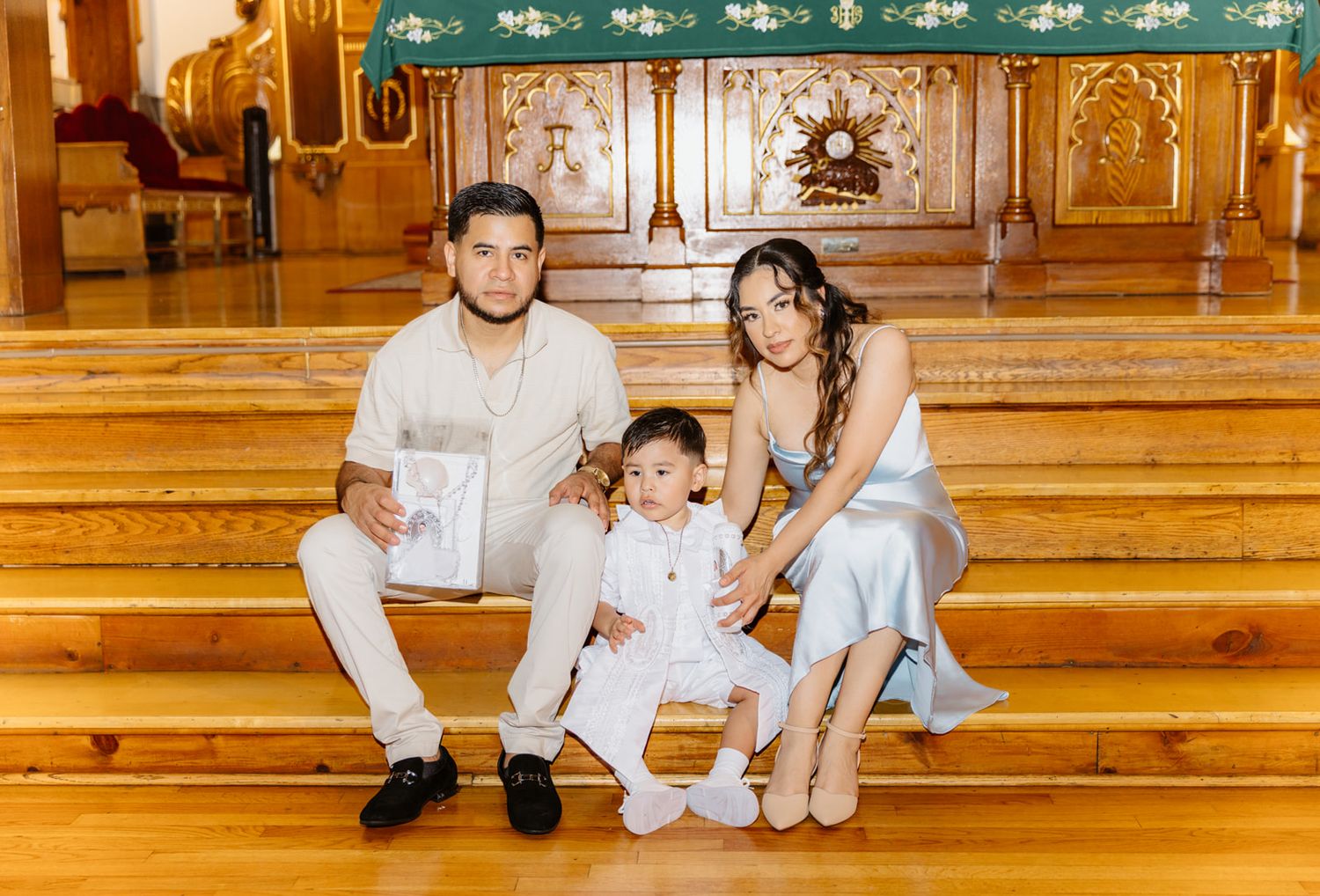 Family sits on church steps during baptism, father holding certificate, child in white dress.