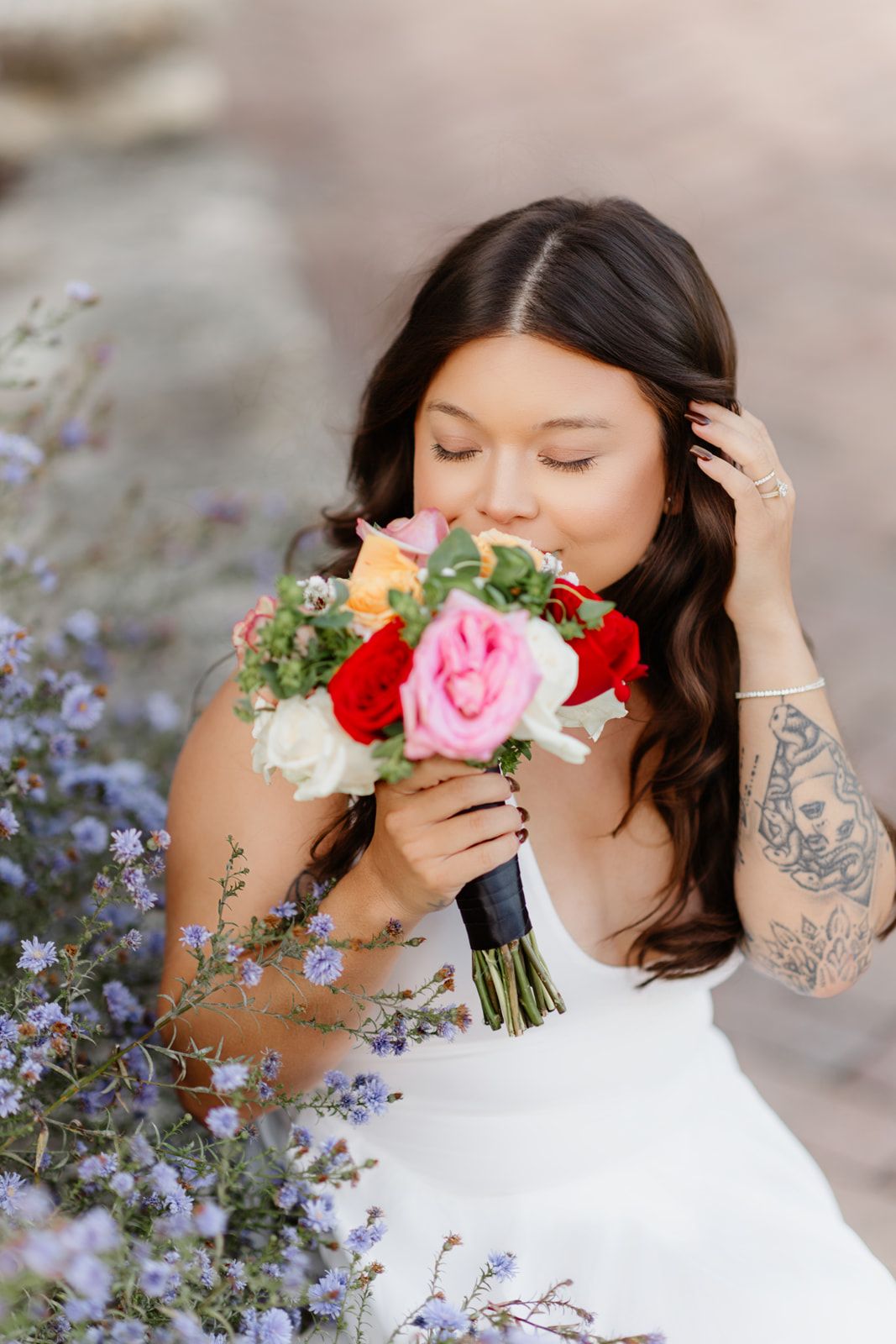 Bride smelling colorful wedding bouquet with red, pink, and white roses, surrounded by purple wildflowers.