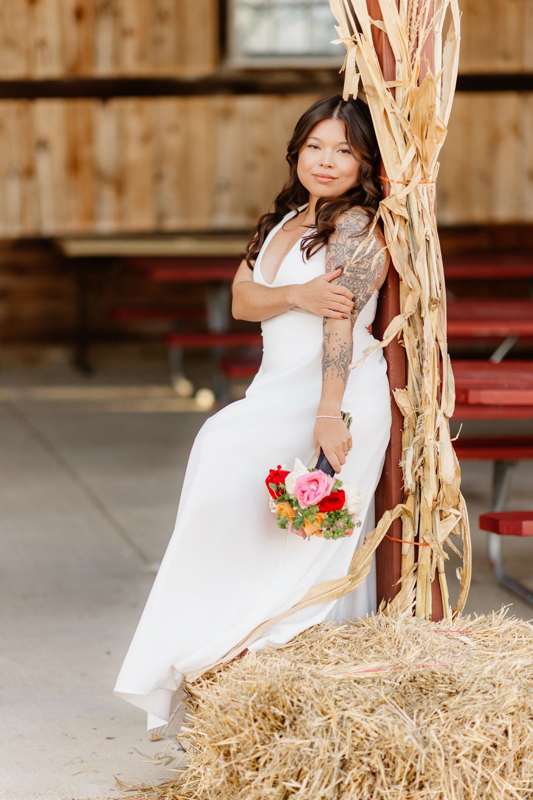 Bride in white gown with floral bouquet poses on hay bale near dried corn stalks in rustic barn setting.