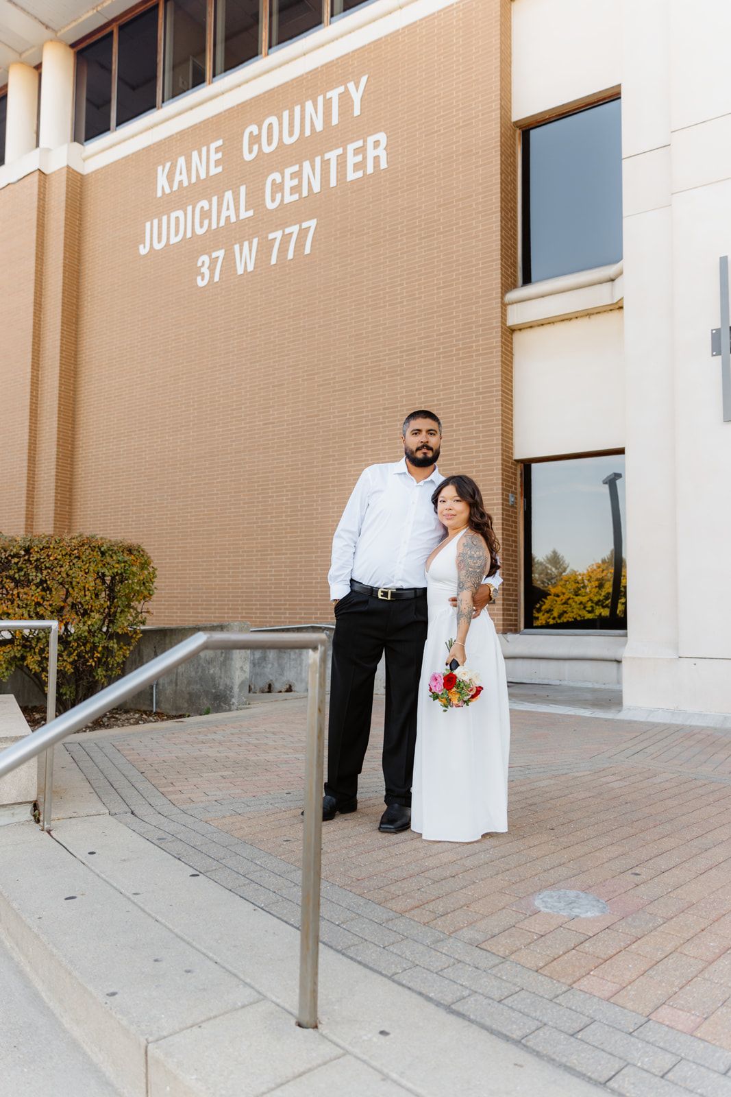 Newlywed couple posing outside Kane County Judicial Center, bride in white gown with bouquet, groom in white shirt.