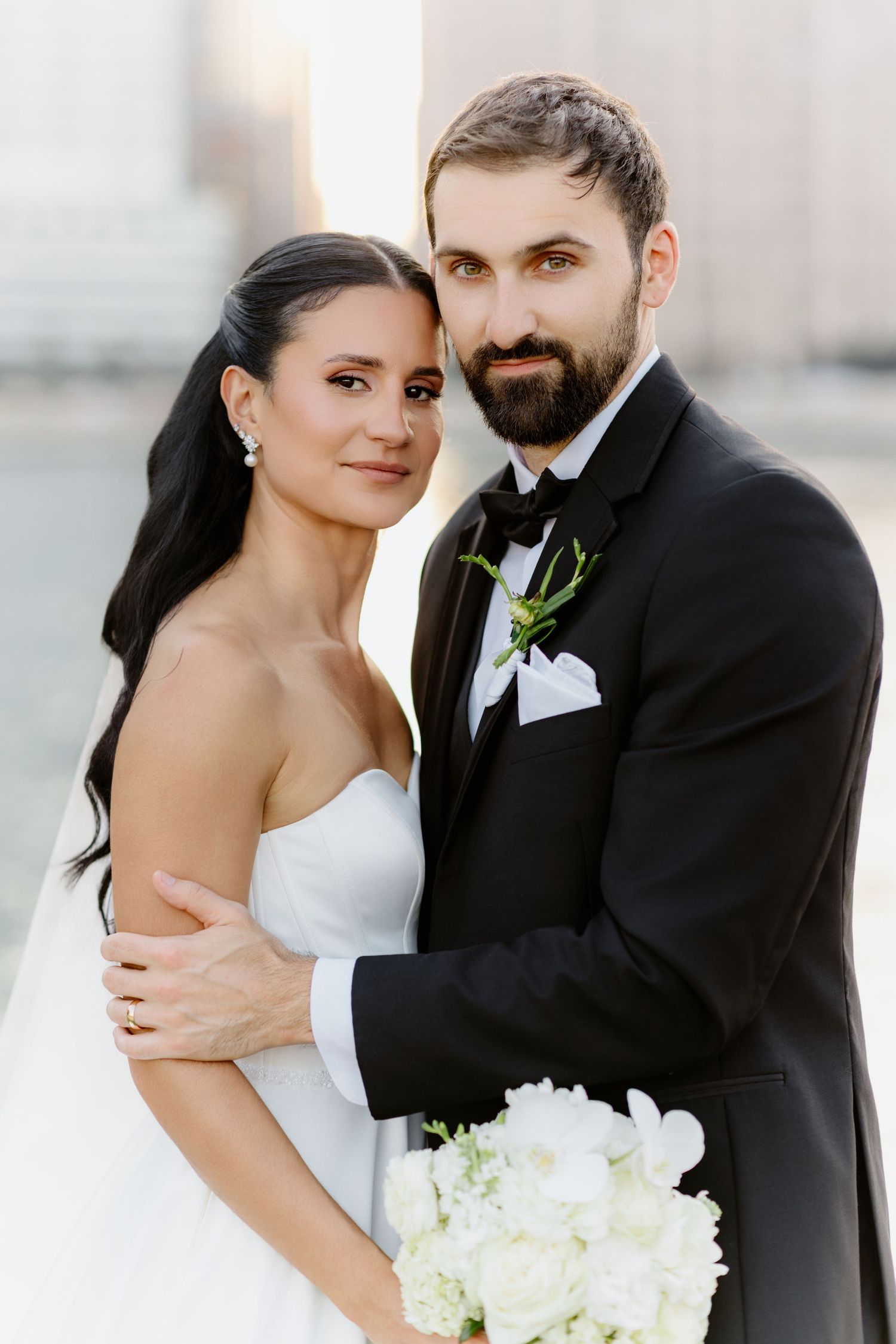 Bride and bearded groom in black tuxedo posing together holding white bouquet.