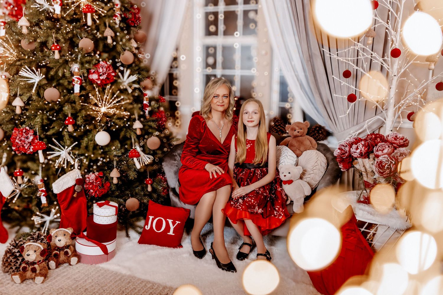 Woman and girl in matching red dresses pose beside decorated Christmas tree in festive holiday room.