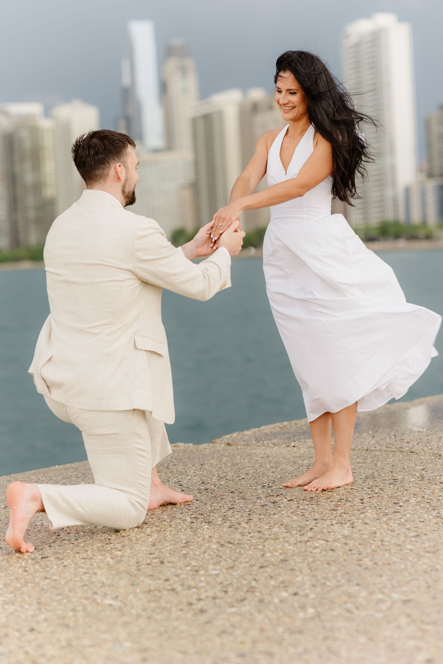 Joyful couple during marriage proposal by Chicago lakefront, man on one knee holding her hands.