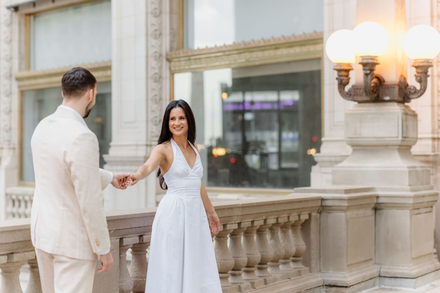 Bride in white halter dress holds groom's hand on ornate balcony with elegant lamp posts in Chicago.
