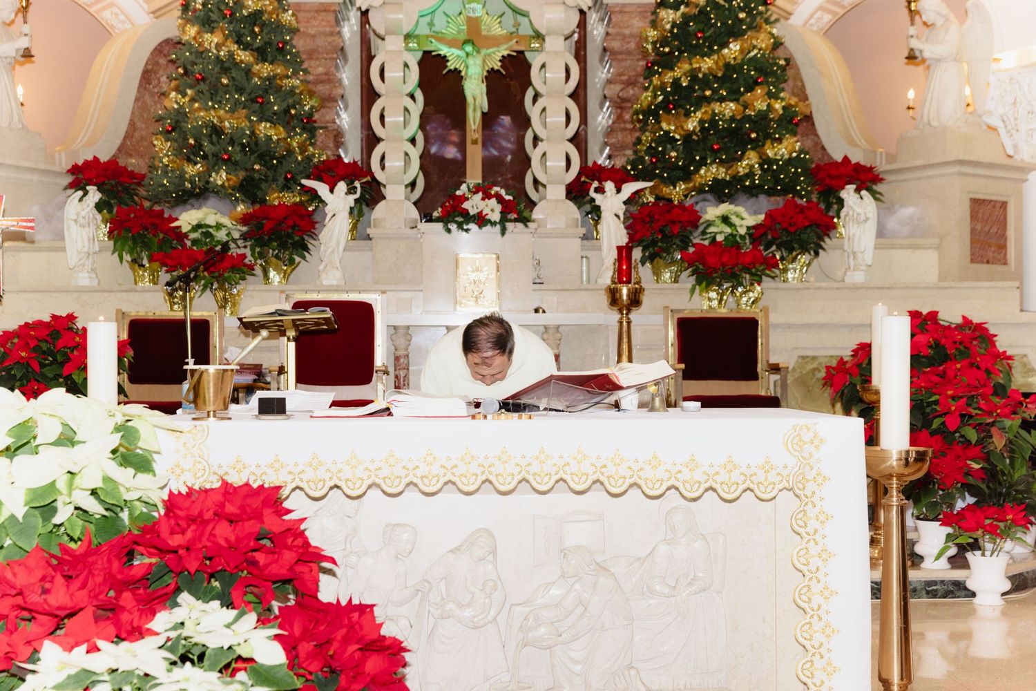 Priest reading at altar surrounded by Christmas trees and red poinsettias during holiday wedding.