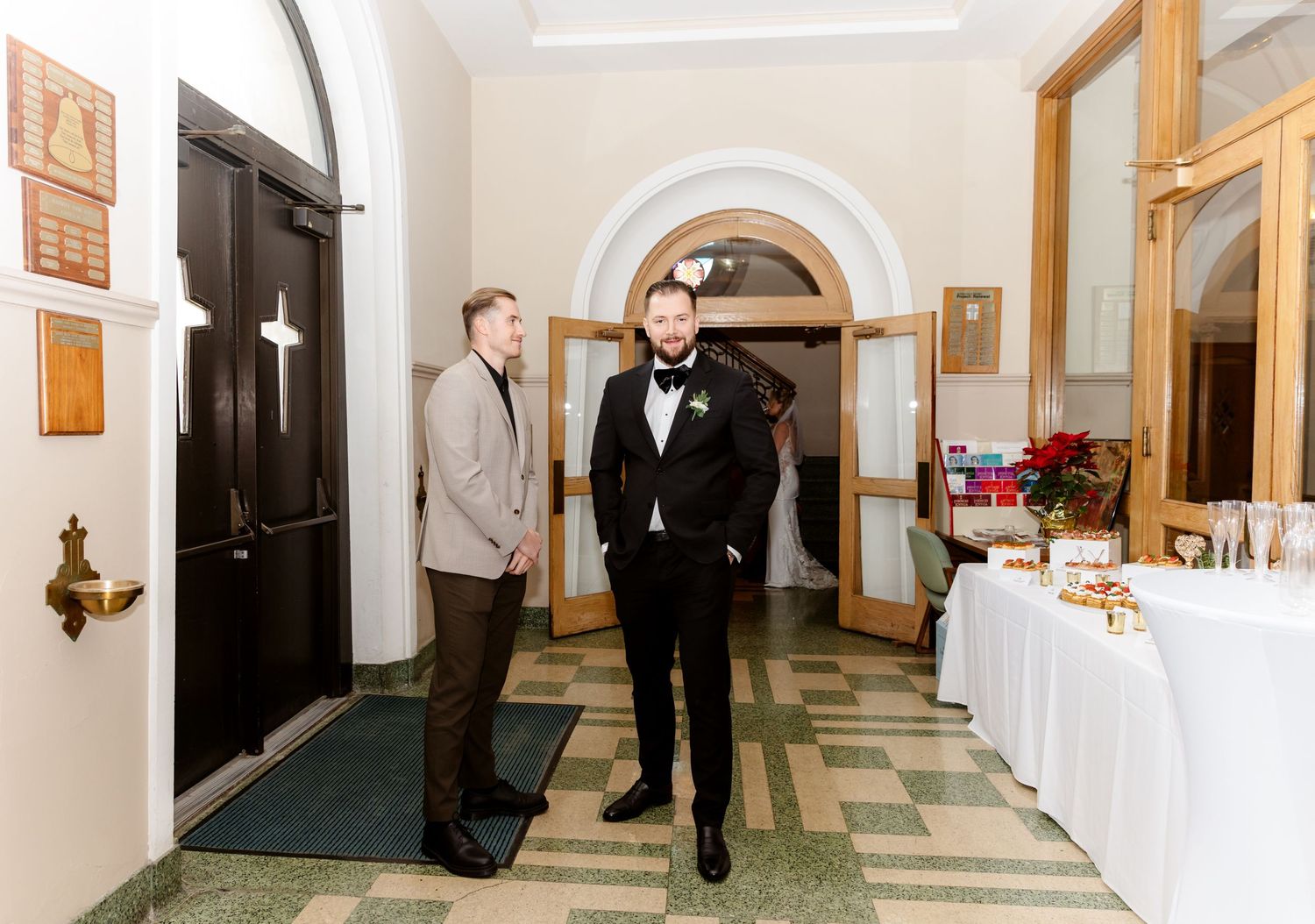 Two men in formal attire standing in a church entrance foyer near a decorated table.