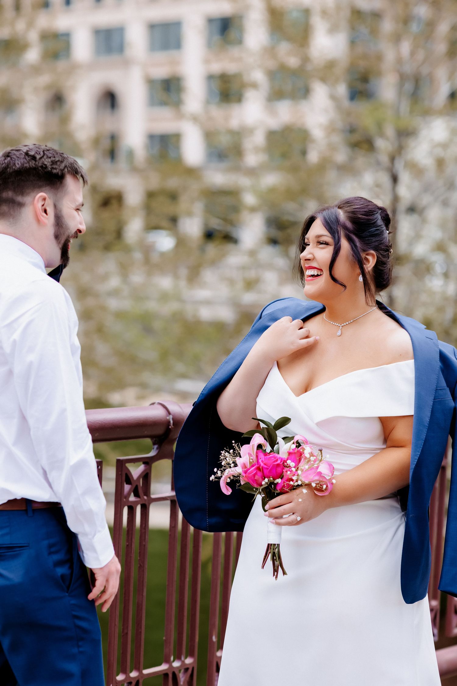 Radiant bride in white dress drapes blue jacket over shoulder and smiles at groom on decorative urban bridge.