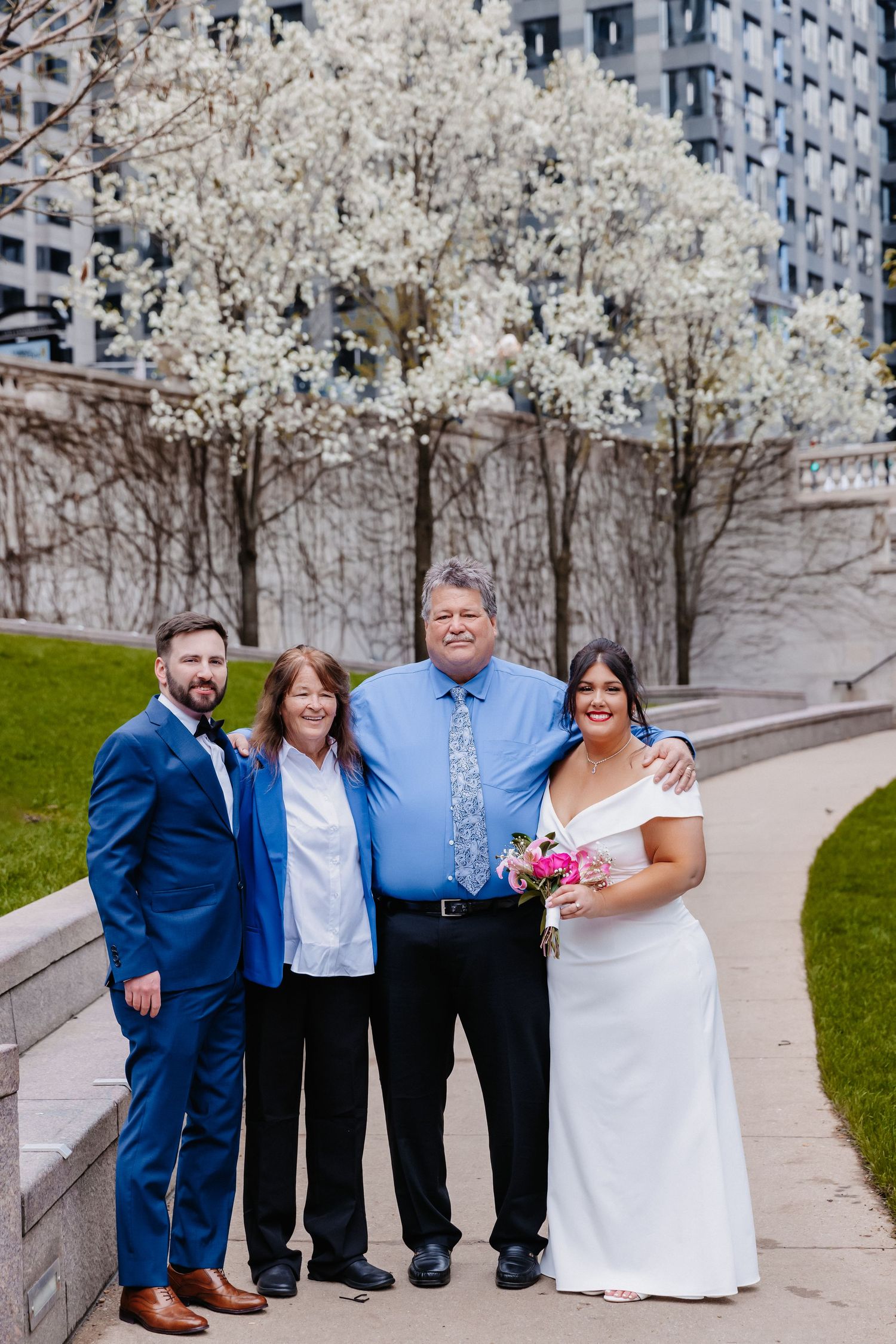 Four wedding guests smile together outdoors near flowering trees with city buildings in background.