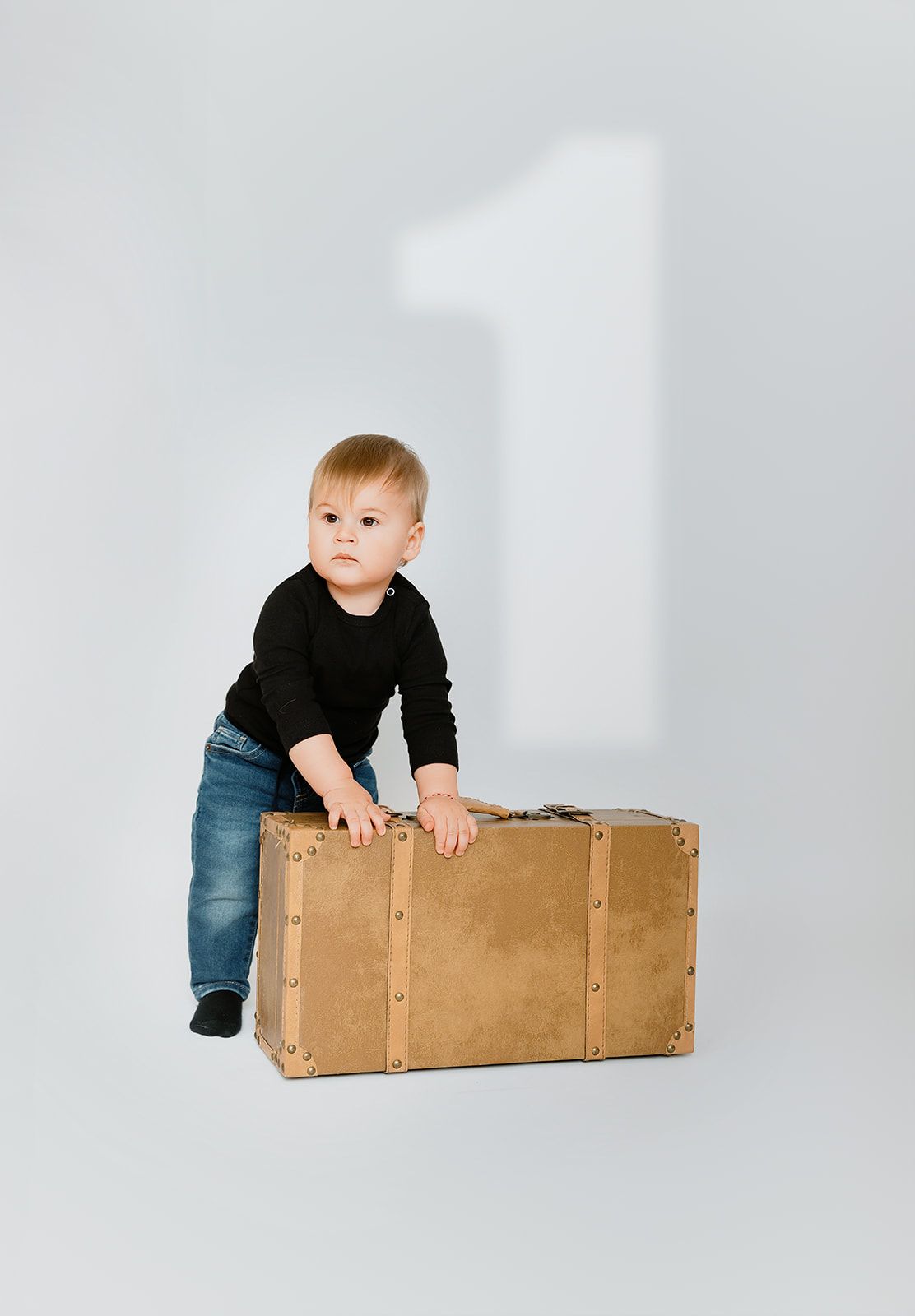 A toddler in a black sweater and jeans stands behind a vintage brown suitcase with a number one on the wall.