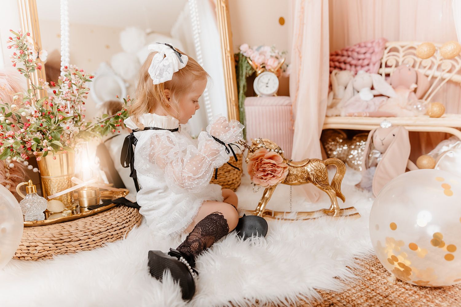 Young girl in white lace dress plays with golden rocking horse in elegant, balloon-filled holiday room.