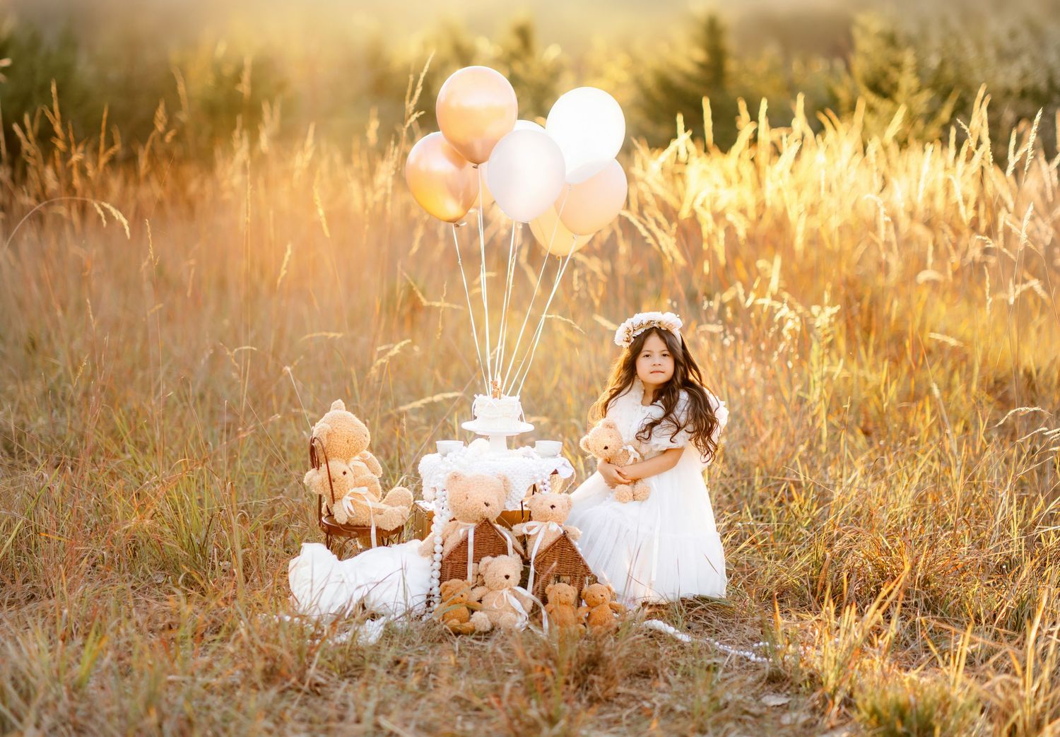 Girl in white dress sits in golden field with balloons and cake for a dreamy birthday photo session.