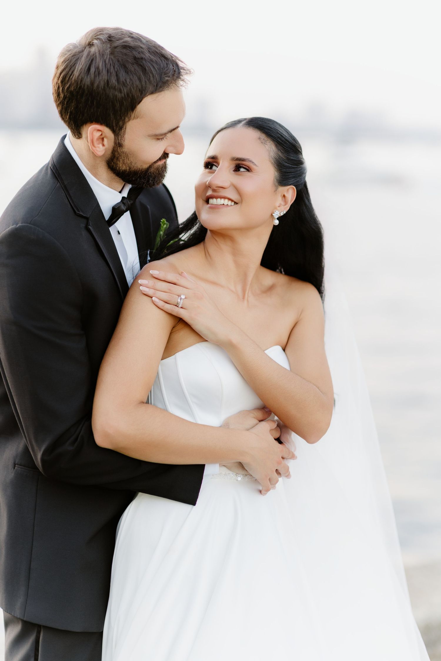 Groom in black tuxedo embraces bride in strapless white gown from behind as she gazes up at him lovingly.