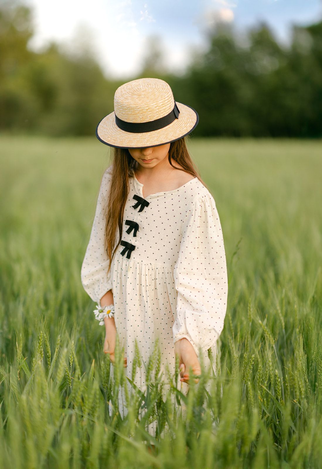 A young girl in a cream polka dot dress and straw hat walks through a lush green wheat field.