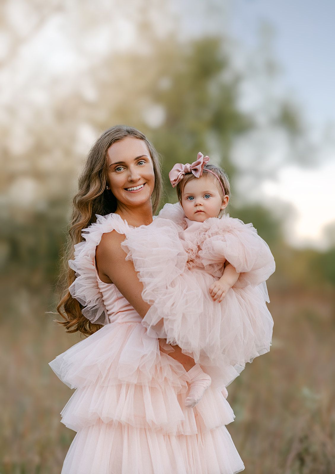A mother in a pink ruffle dress holds her baby girl in a matching pink tutu dress outdoors.