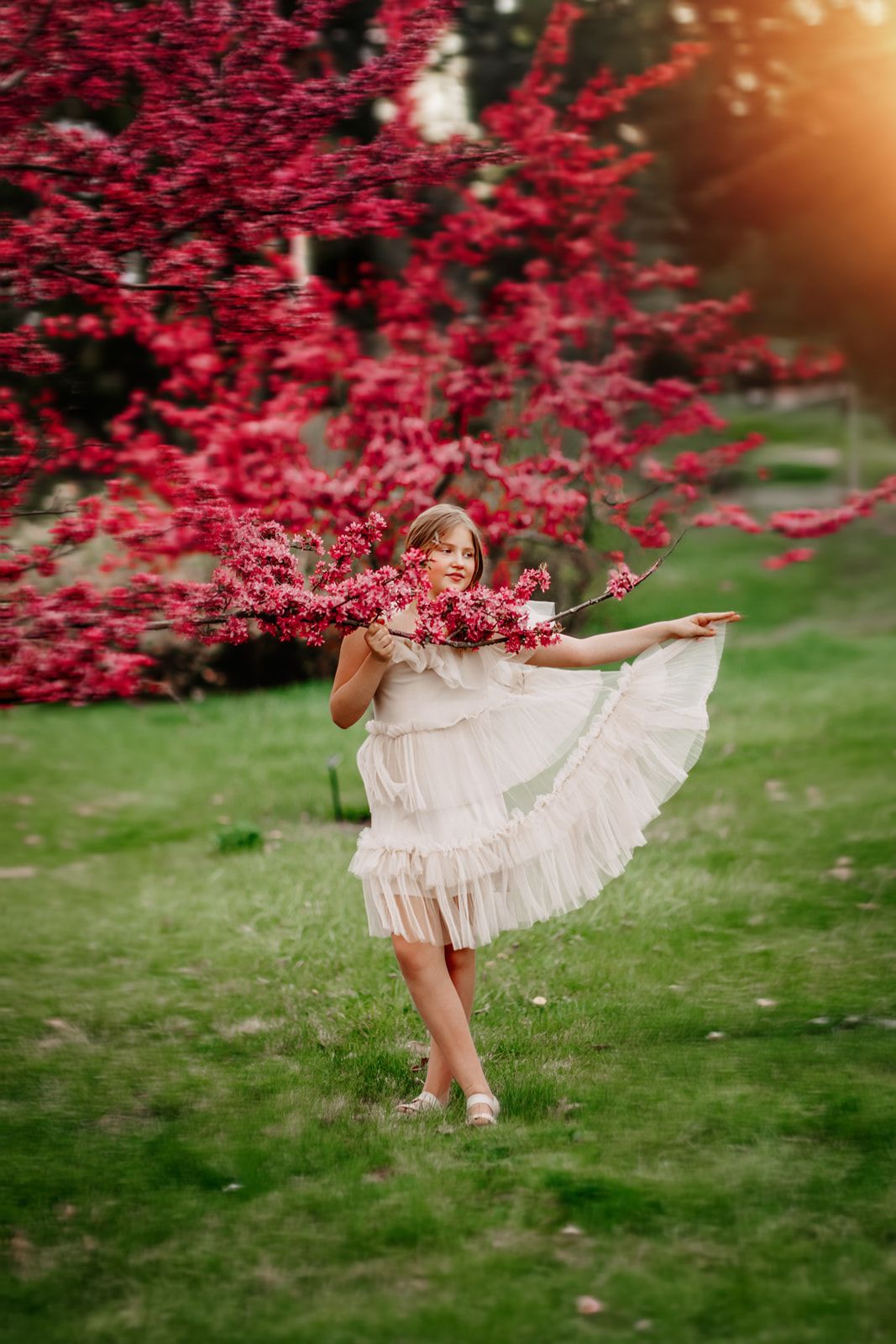 Young girl in white ruffled dress dancing near vibrant red flowering tree on green grass in golden sunlight.