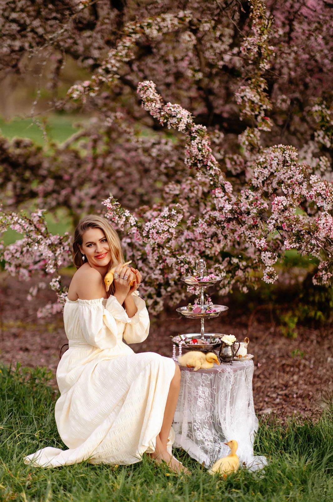 Woman in white off-shoulder dress sitting by blooming cherry tree with tiered dessert stand outdoors.