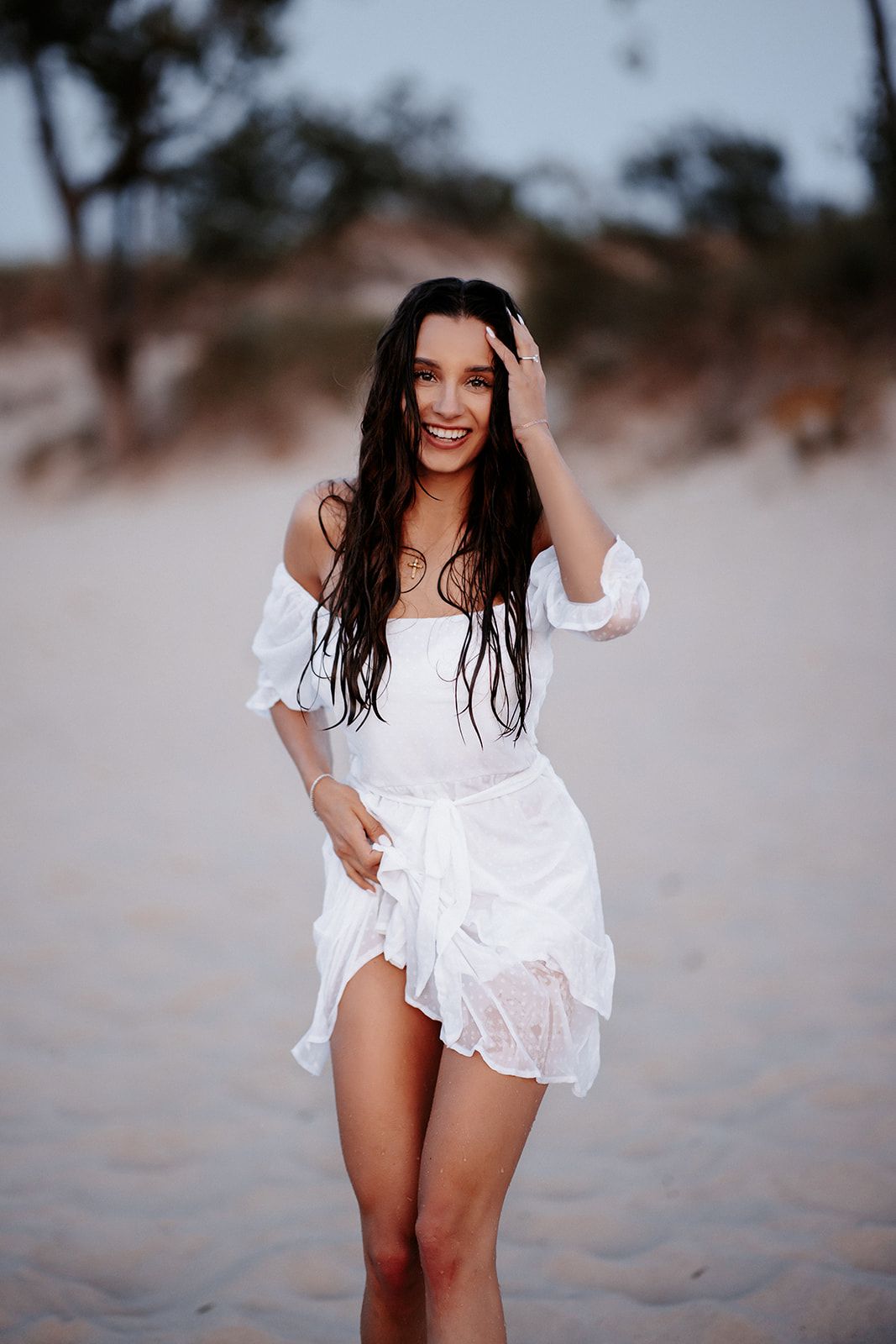 Smiling brunette woman in white off-shoulder dress posing on sandy beach with wet hair and trees in background.