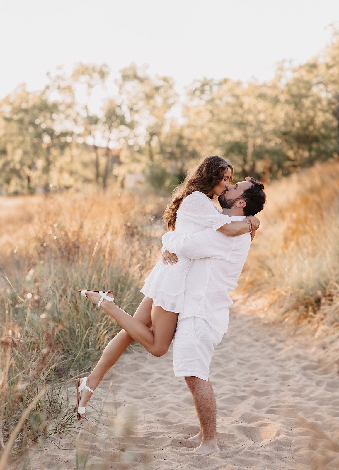 Couple in white outfits sharing a romantic kiss on sandy path, man lifting woman at golden hour.