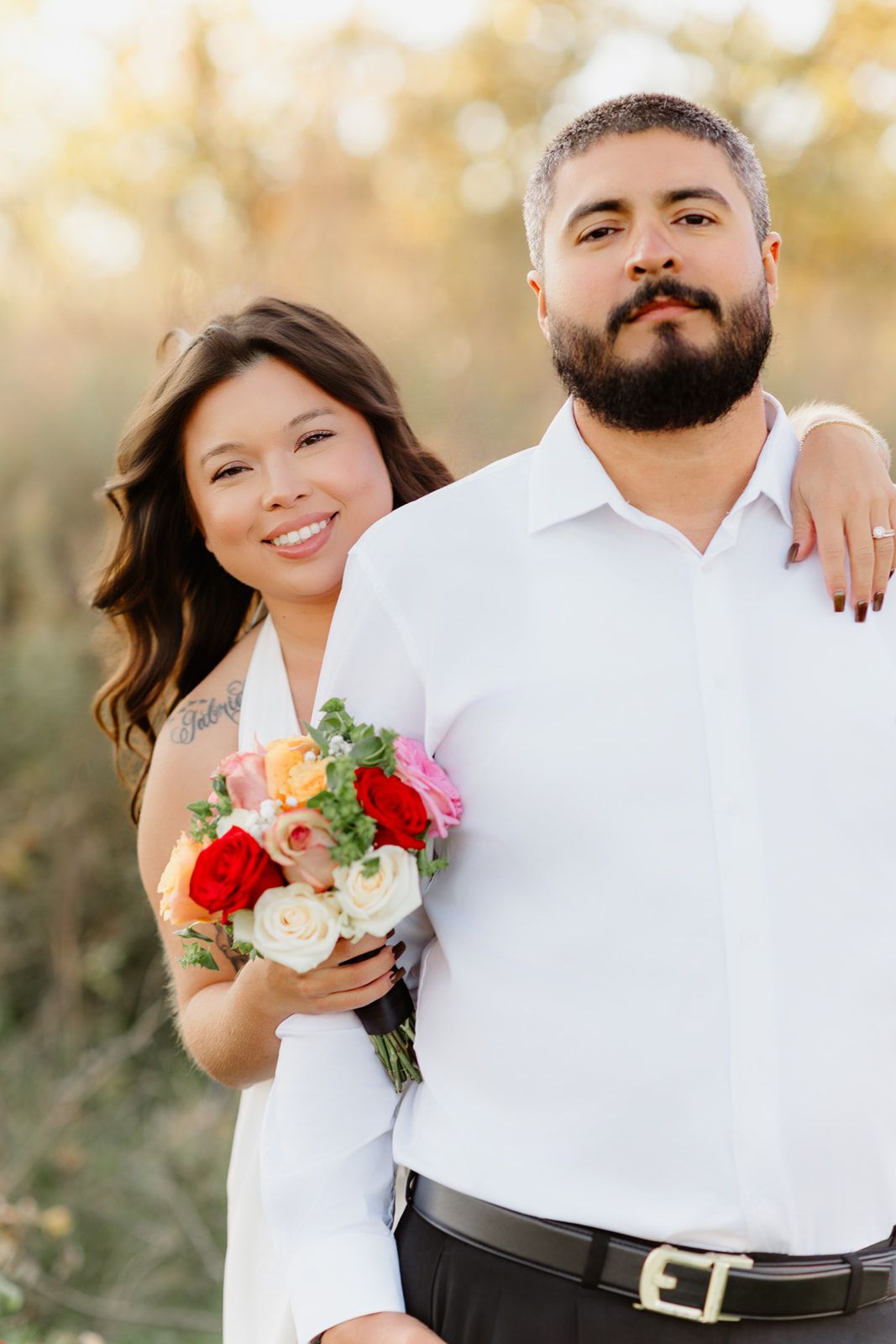 Bride holding colorful bouquet smiles behind bearded groom in white shirt outdoors.