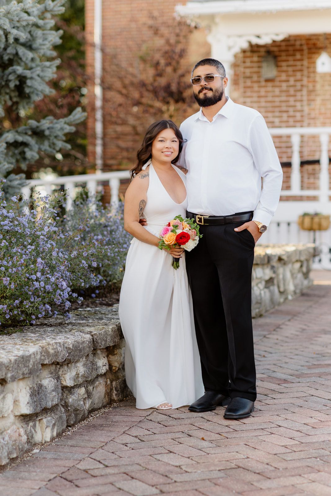 Bride in white dress holding colorful bouquet stands with groom in white shirt and black pants on brick path.