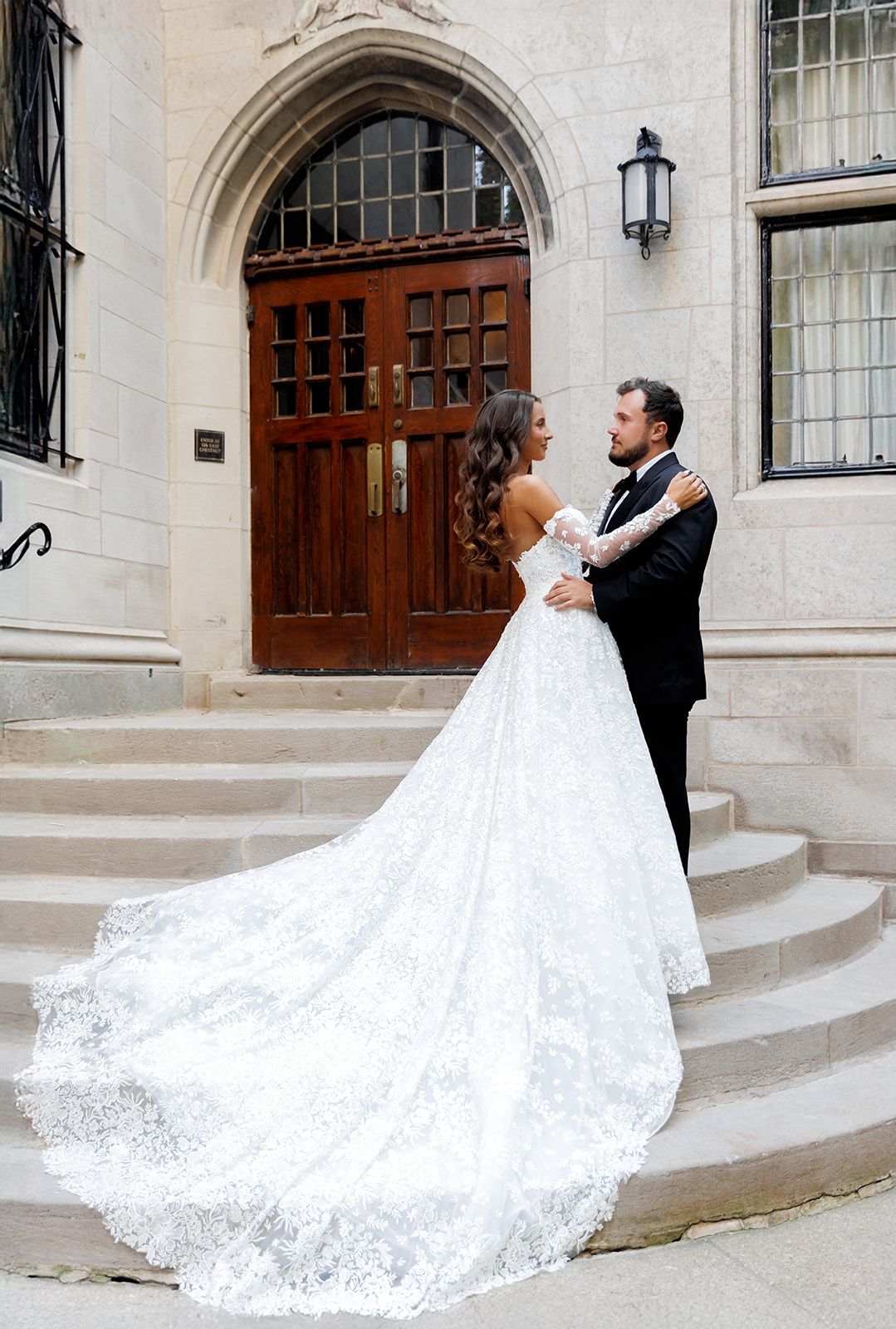 Bride in lace ball gown with long train stands with groom on stone steps before elegant Gothic arched wooden doors.