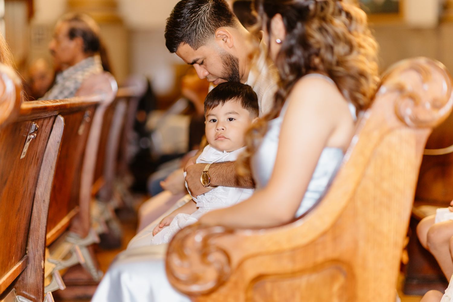 A family sits in wooden church pews during a ceremony, with a father tenderly kissing his toddler dressed in white.