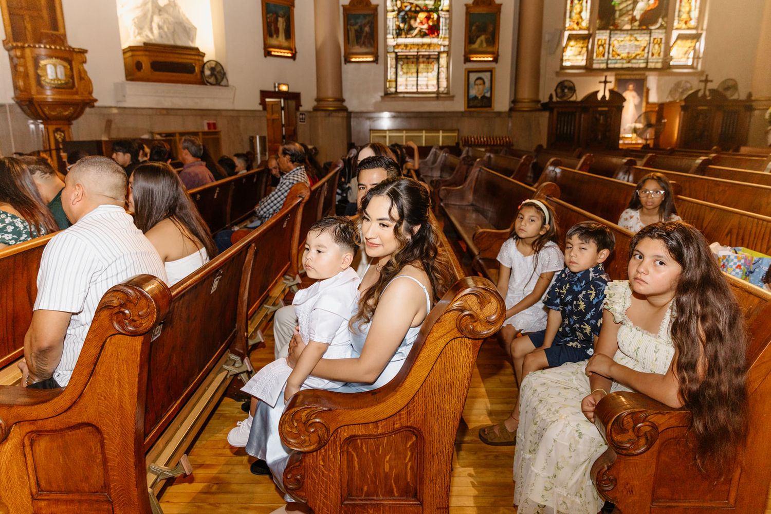 Families with children seated in wooden church pews during a Catholic baptism ceremony.