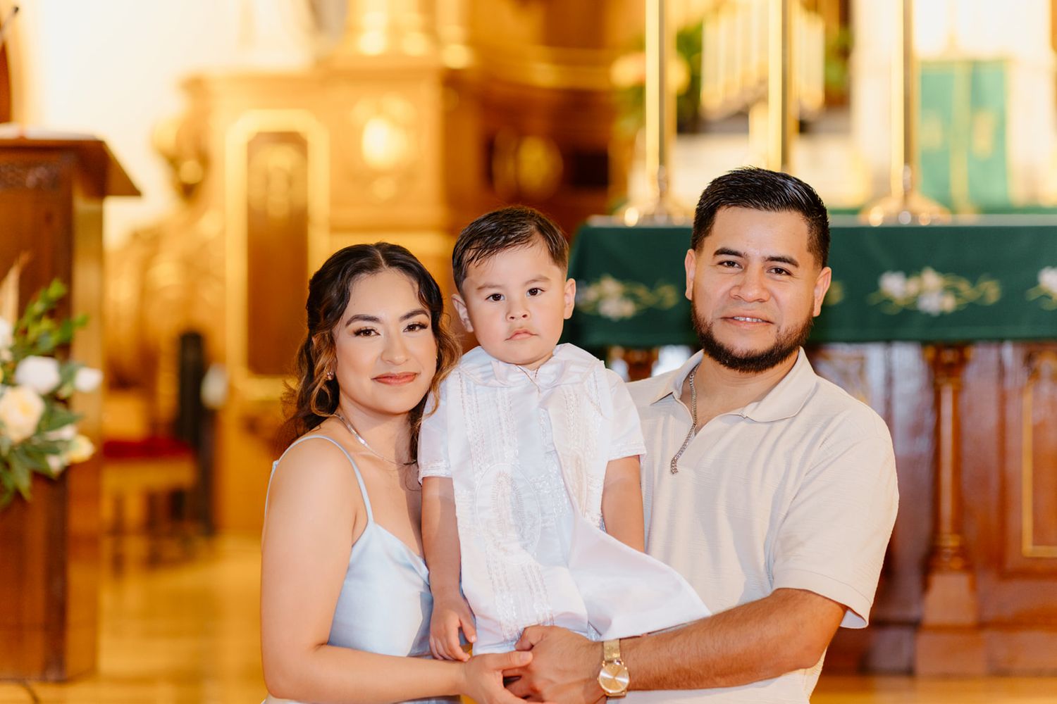 A couple holds their baby dressed in white baptism clothes while posing inside a golden-lit church.