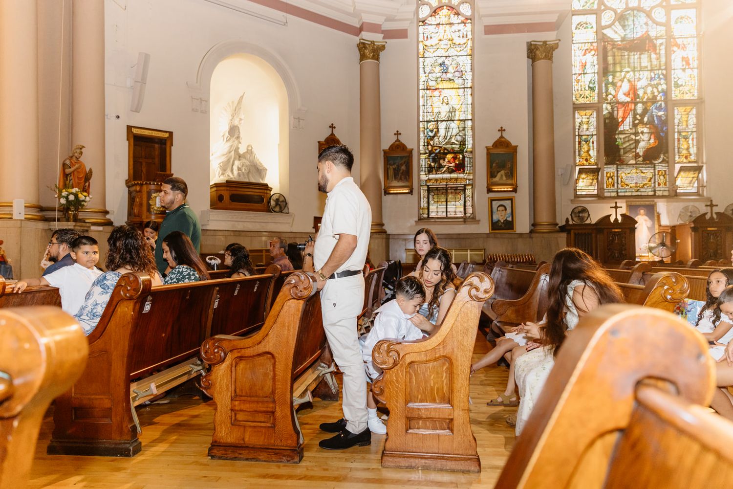 Children in white attire sit in wooden church pews during a religious ceremony with stained glass windows.