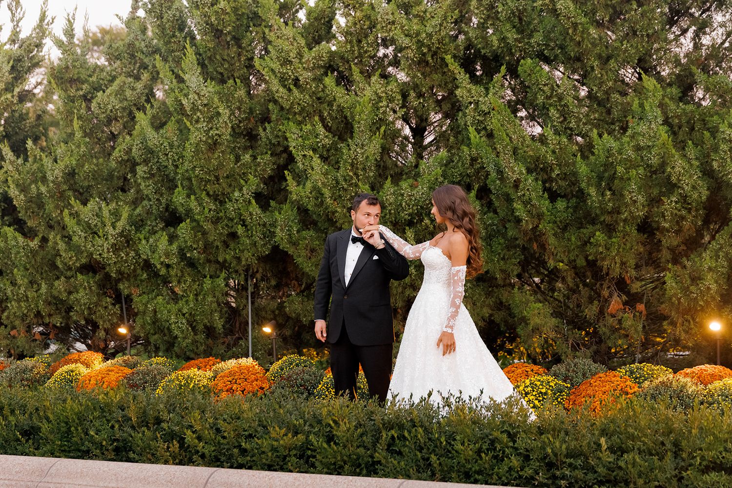 Bride and groom share a romantic moment outdoors among autumn mums and evergreen trees at their fall wedding.