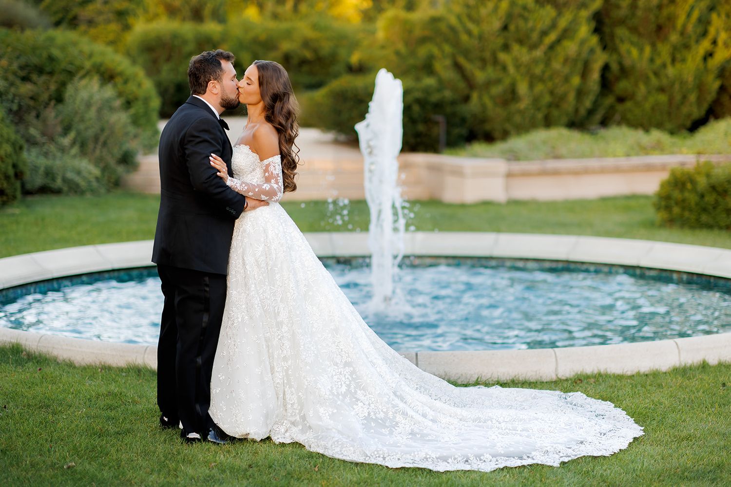 Bride and groom share a romantic kiss in front of a garden fountain, bride wearing a lace ball gown with long train.