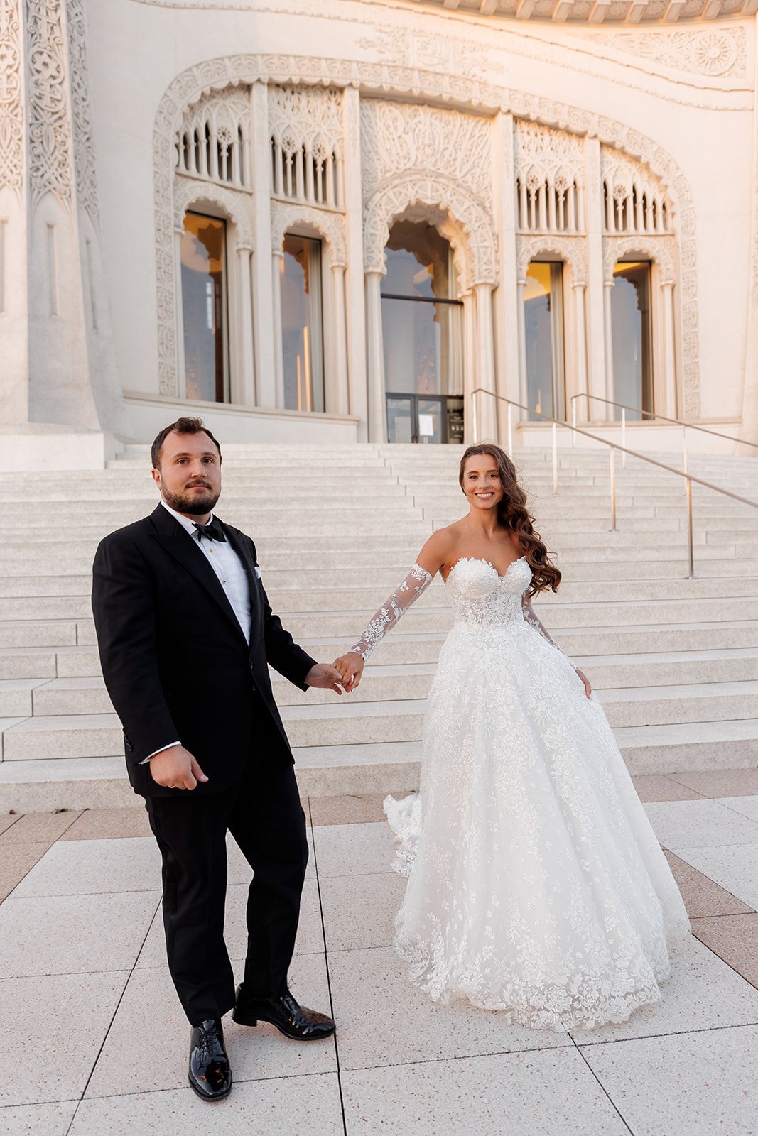 Smiling bride in lace ball gown and groom in tuxedo hold hands on steps of ornate building.