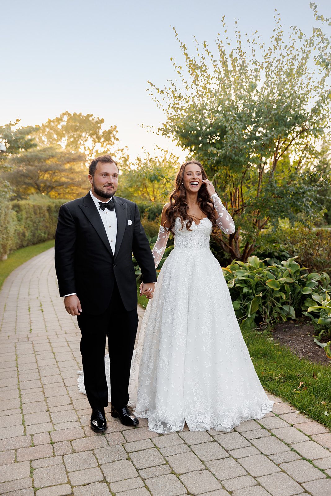 Smiling bride in lace gown holds hands with tuxedo-clad groom while walking along a garden brick path at sunset.