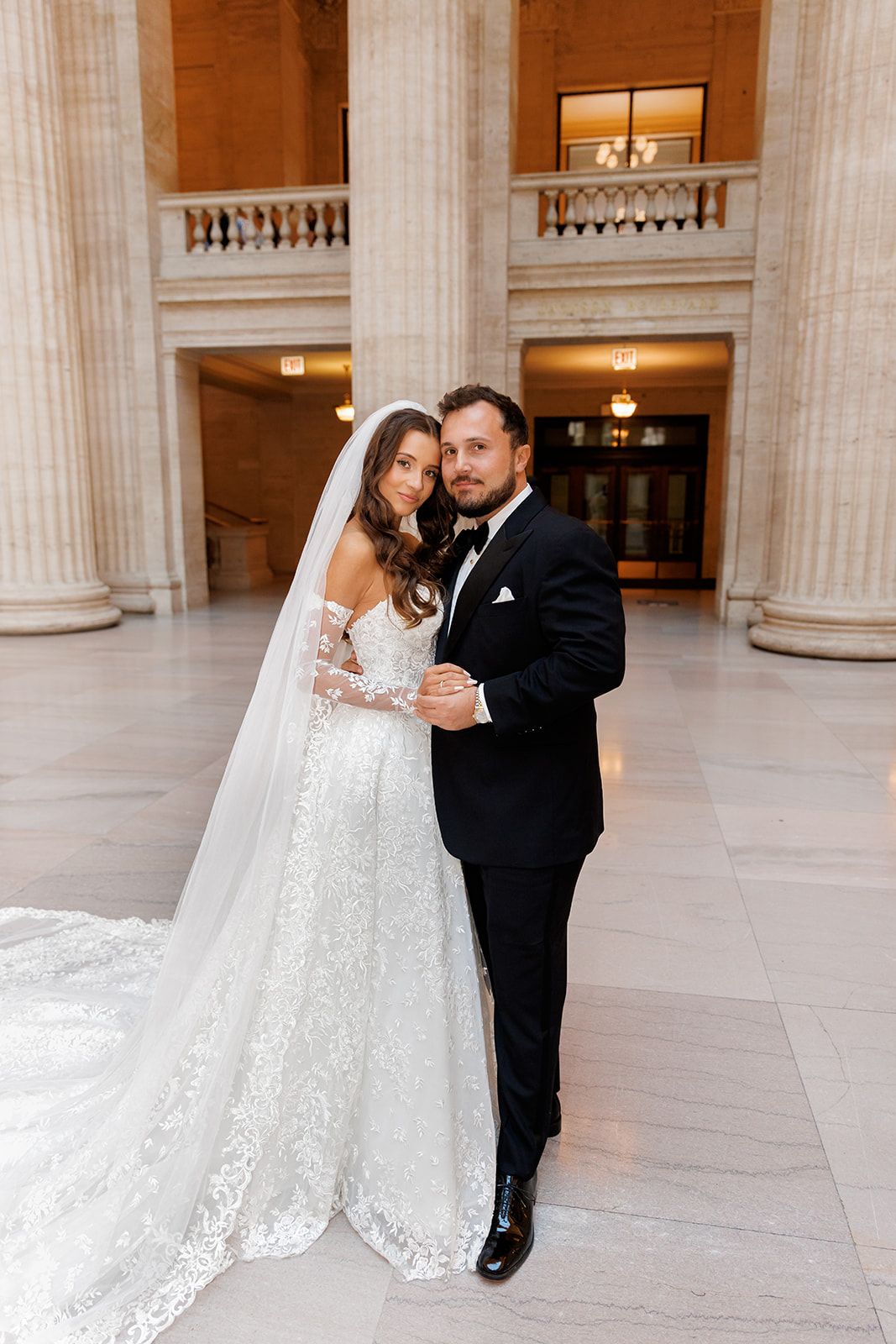 Bride and groom pose elegantly in a grand marble hall with tall columns, she wearing a lace gown with long veil.