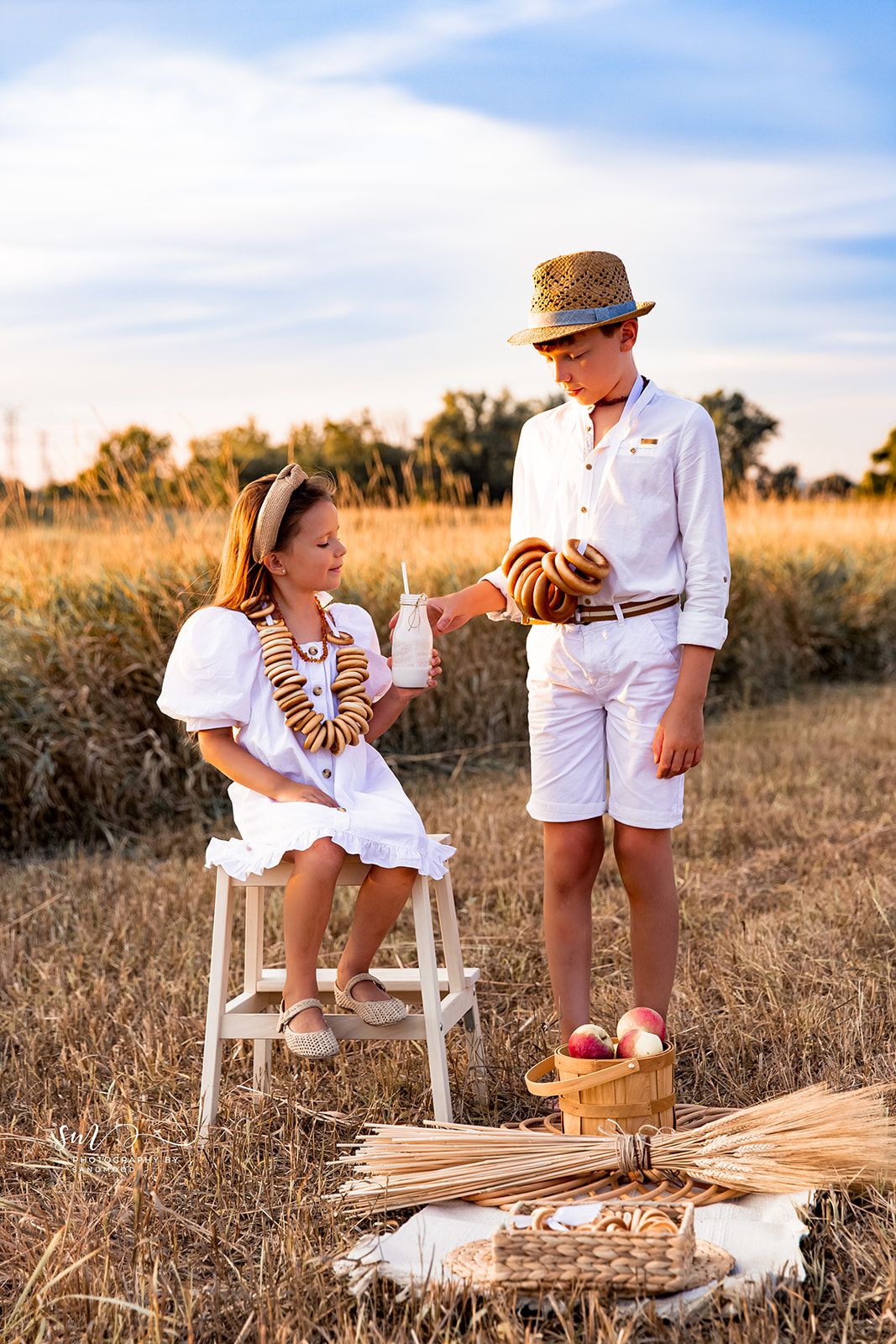 Two children in white outfits enjoy a picnic in a golden wheat field at sunset, sharing bread and milk.