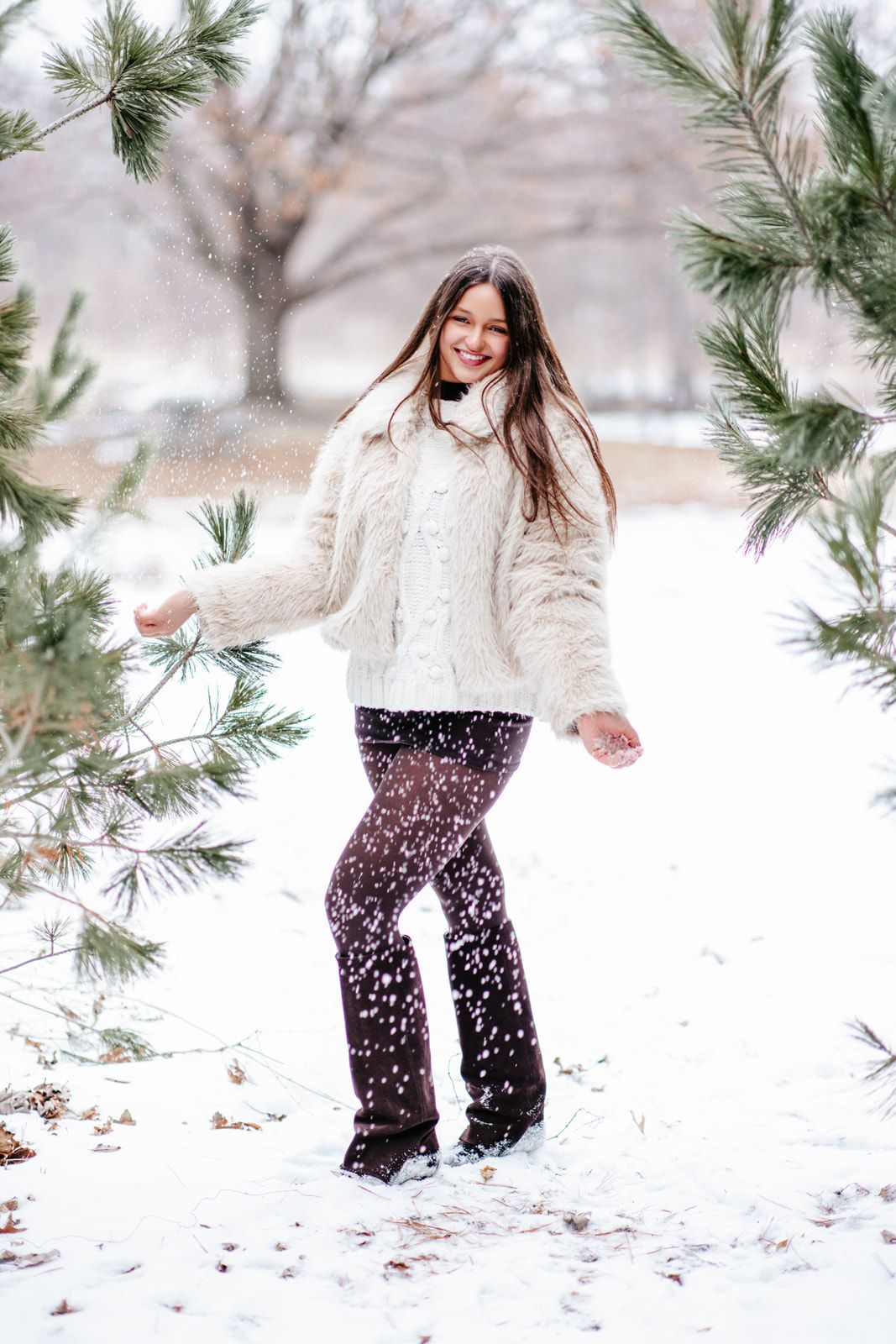 Young woman in white fur jacket playing in snow among pine trees, smiling joyfully on a winter day.