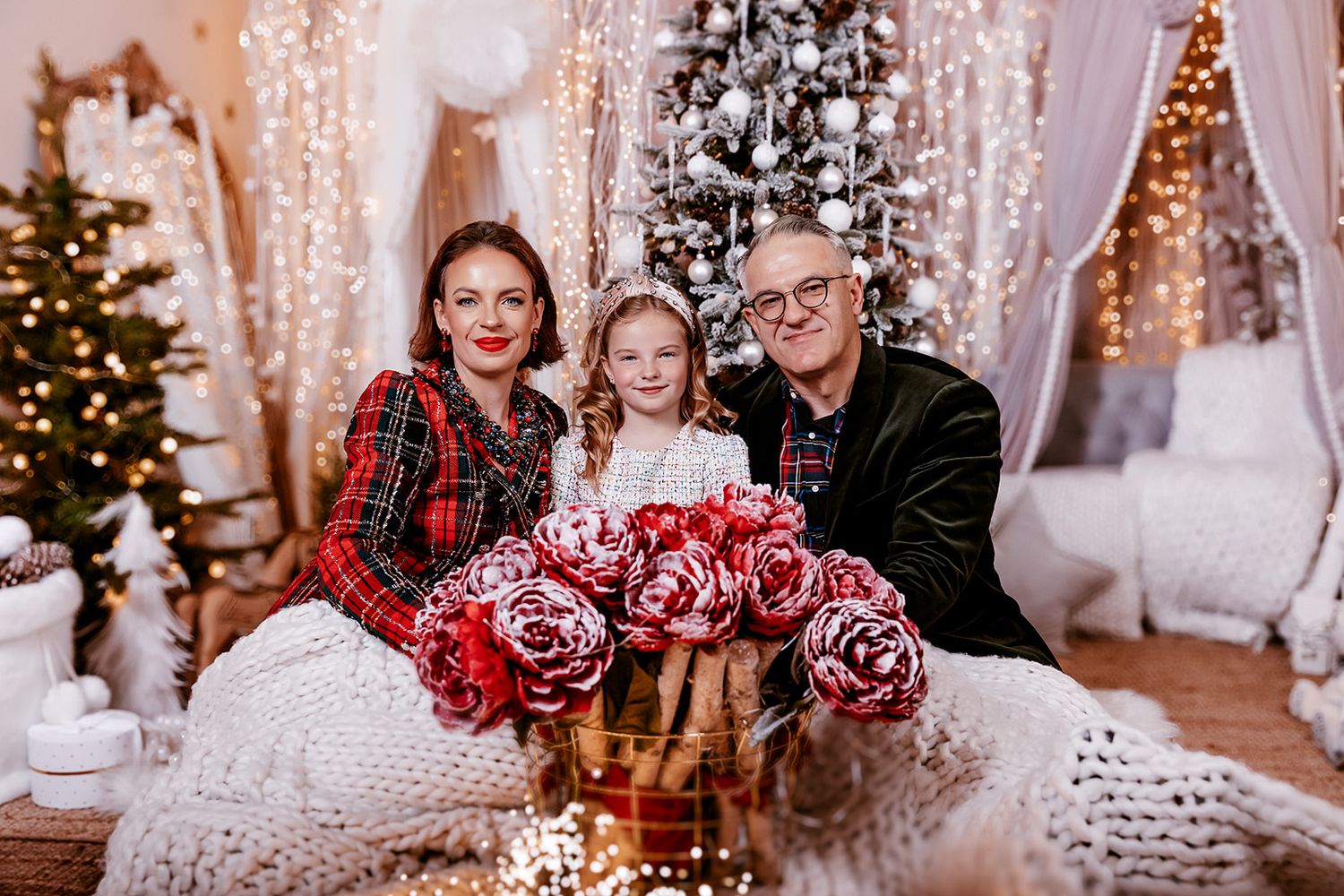 Family of three posing with red roses near a decorated Christmas tree with warm holiday lights.