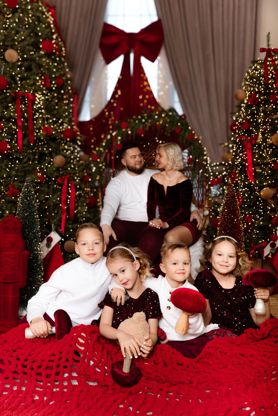 Family Christmas photo with four children on red blanket and parents near decorated trees with red bows and lights.