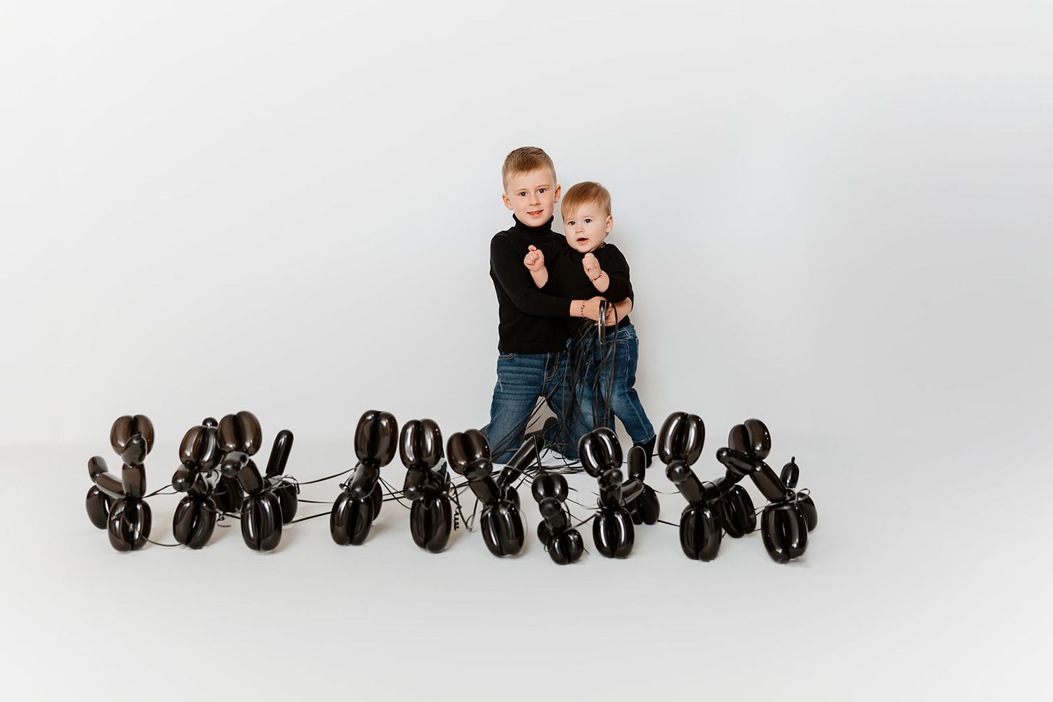 Two children in black outfits pose surrounded by shiny black balloon animal dogs on a white background.