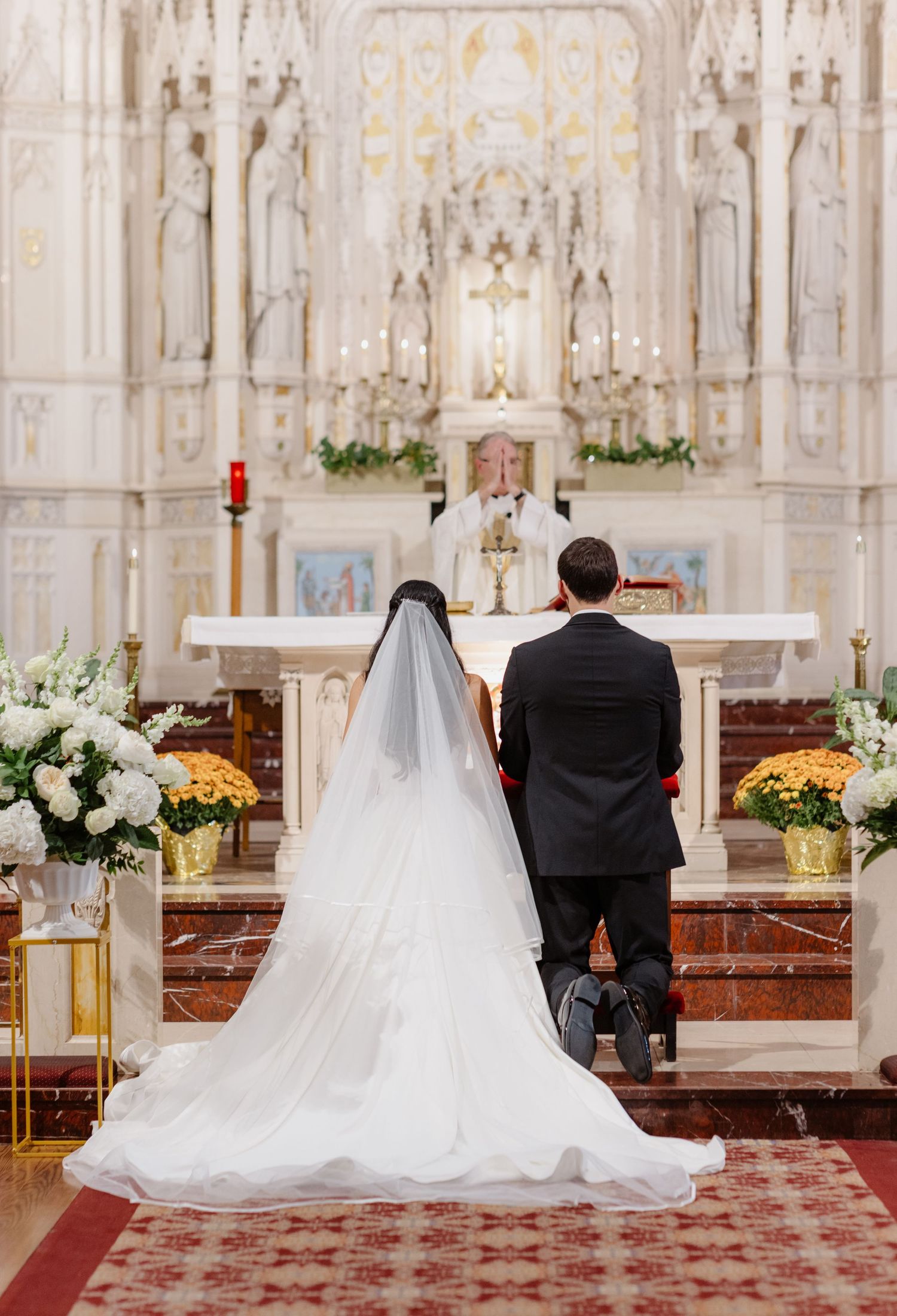 Bride with long veil and groom kneel at ornate white church altar.