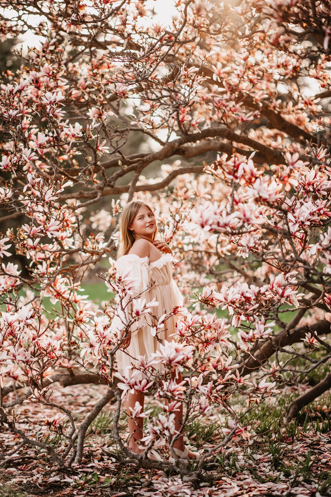 Young woman in white dress standing among blooming pink magnolia tree branches in spring garden.